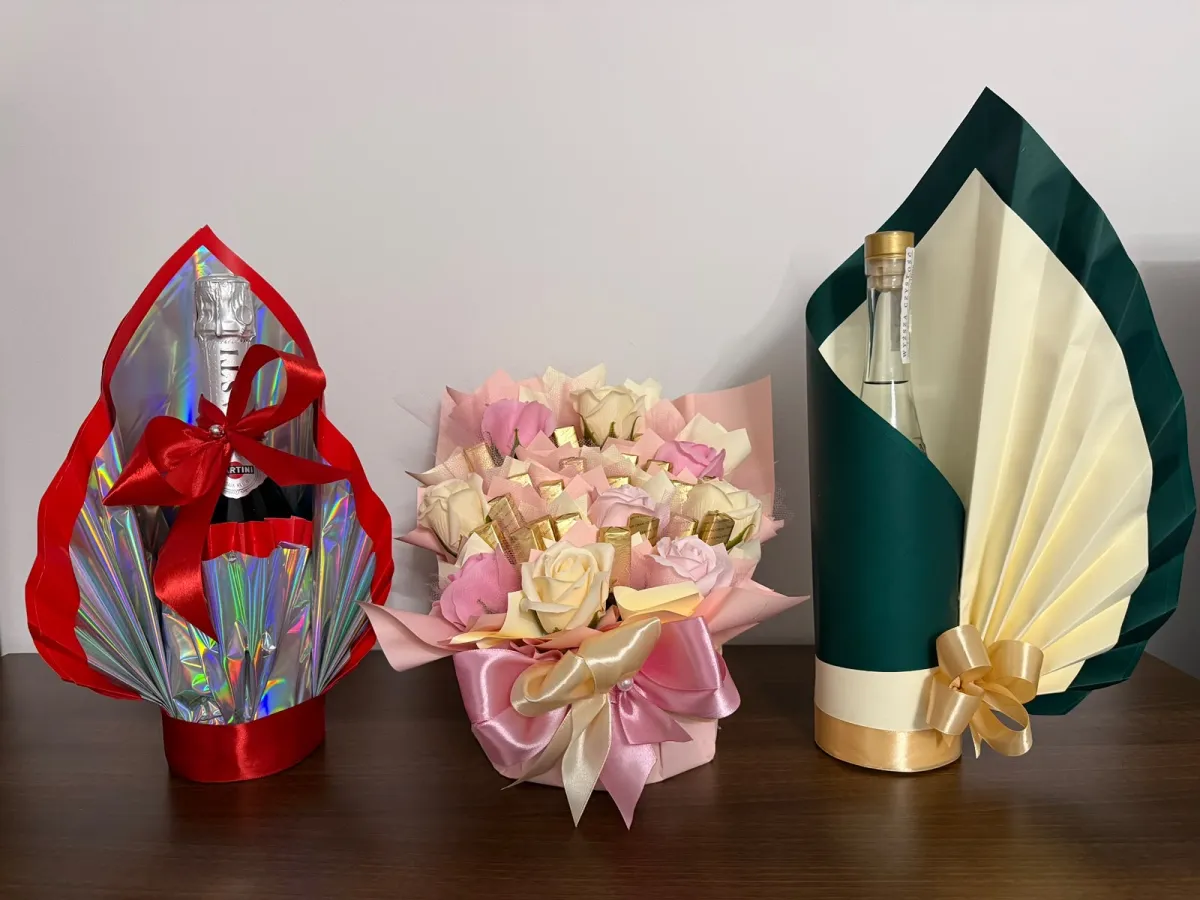 Dwie butelki w plisowanych, dekoracyjnych oprawach — czerwono-holograficznej i zielono-kremowej — oraz mały bukiet z różami i czekoladkami